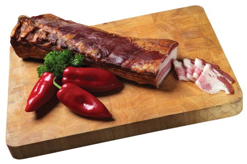 A piece of dry-aged Mangalica bacon placed on a wooden board. Sliced bacon, red peppers, and parsley are arranged beside it, with light smoke visible above the product.